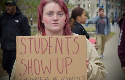 College-aged woman holding a cardboard sign that reads \"Students Show Up, So Where is Admin?\"