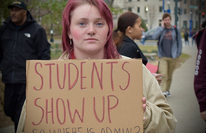 College-aged woman holding a cardboard sign that reads \"Students Show Up, So Where is Admin?\"