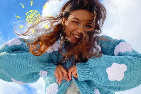 Girl is a white dress and blue sweater with clouds is looking down towards camera smiling. There is an animated/drawn sun in the sky.