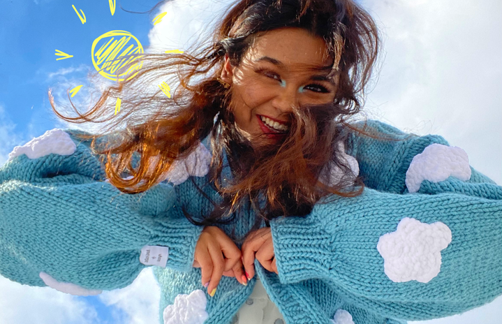 Girl is a white dress and blue sweater with clouds is looking down towards camera smiling. There is an animated/drawn sun in the sky.