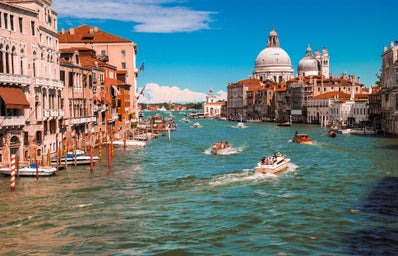 Canal in Venice with multiple boats in the water, with a city skyline in the background.