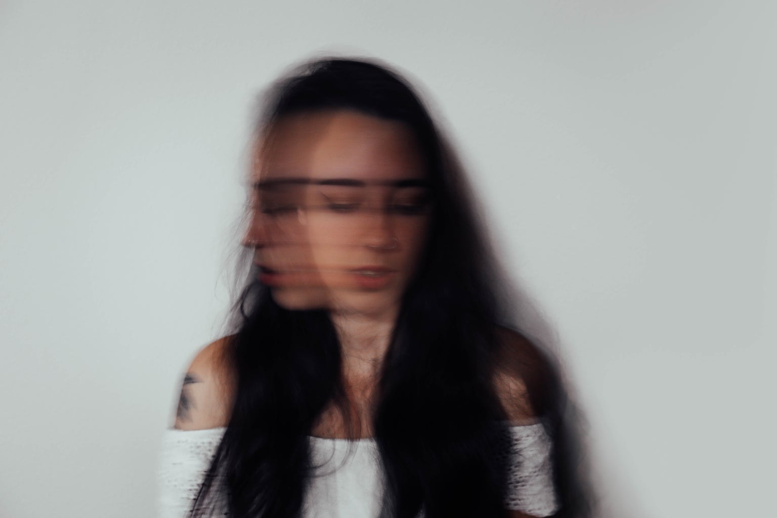 Blurry photo of a woman with dark hair in a white top shaking her head against a grey background.