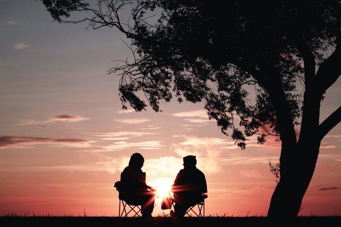 Two silhouettes against a backdrop of an orange-blue sunset are sitting in foldable camping chairs. There\'s a large tree to their right and above them.