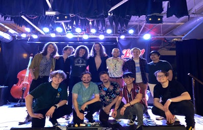 twelve young men standing and kneeling on a stage in rows with instruments and lights behind them