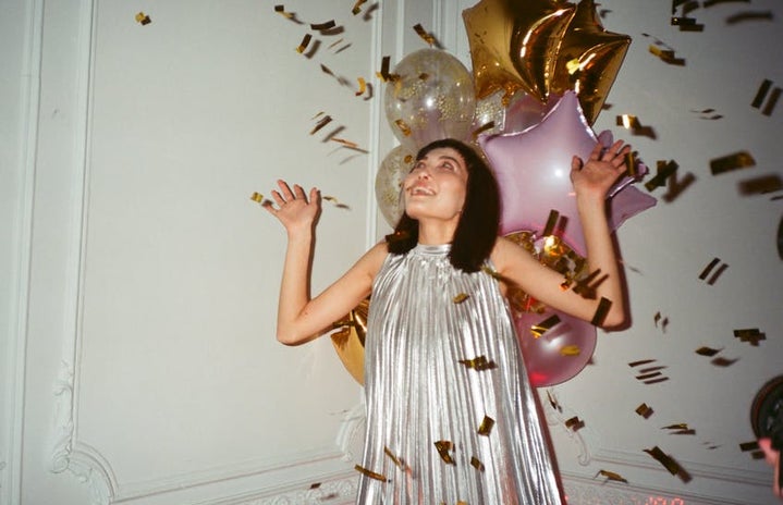 A girl in a silver dress with gold confetti and pink and gold balloons surrounding her