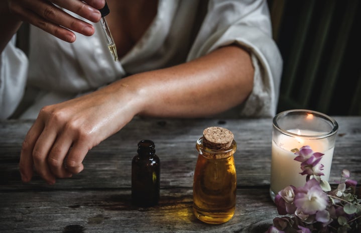 Woman is putting oil on her skin while next to a candle and flowers