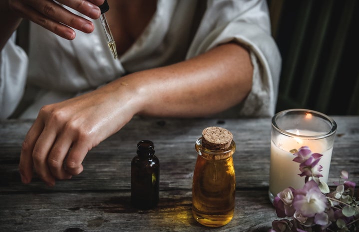 Woman is putting oil on her skin while next to a candle and flowers