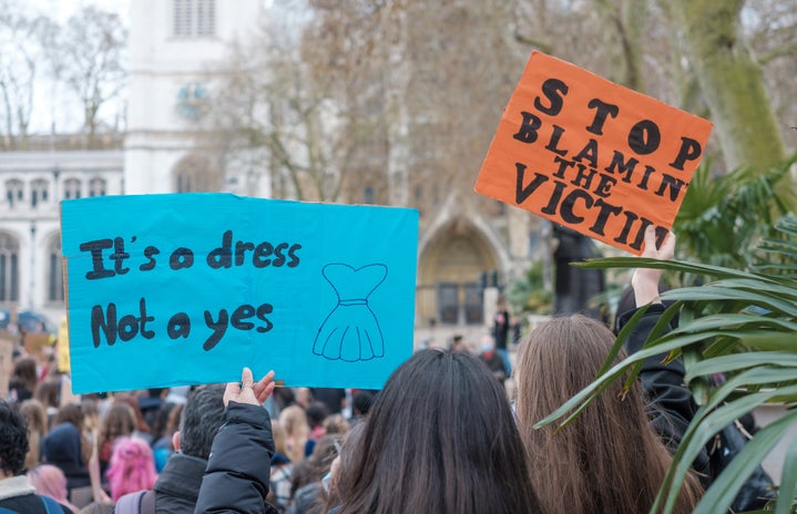 Two signs at a protest. One says \"It\'s a dress not a yes\" and the other \"Stop blaming the victim.\"