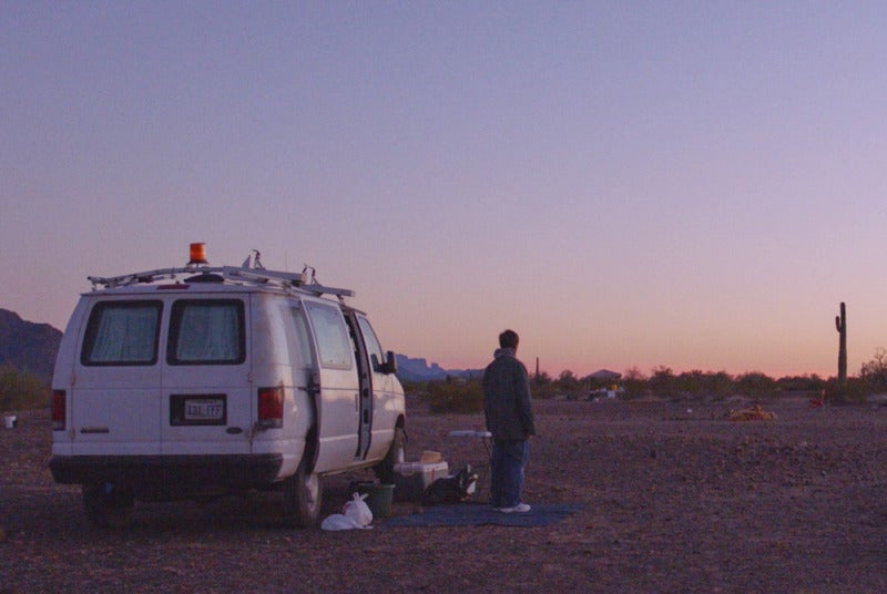 woman standing in front of a white van with her back against the camera, gradient purple sunset in the background
