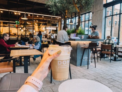 girl holding coffee out with coffee shop seating in the background