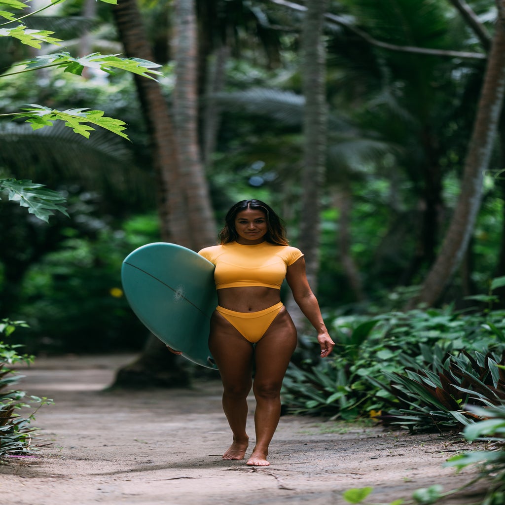 Influencer Eryn Krouse holding a surfboard in the rainforest with trees in the background.