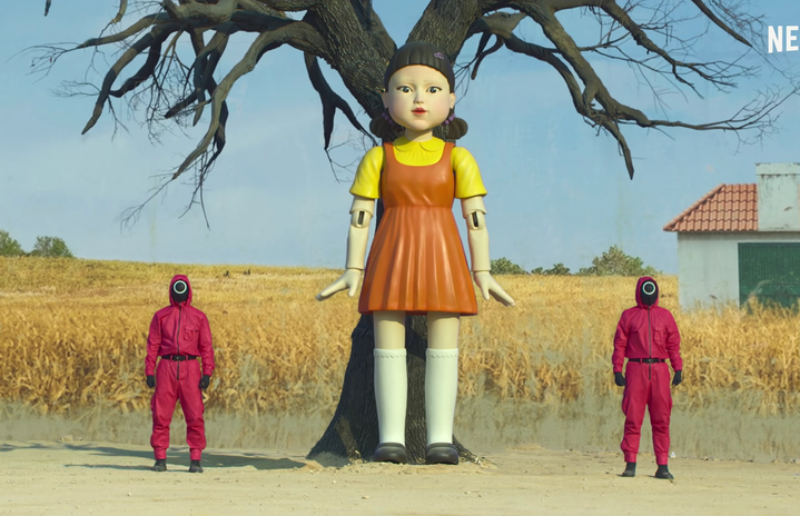 screenshot of a mechanical doll and two workers dressed in red from the Squid Game Official Trailer on YouTube by Netflix