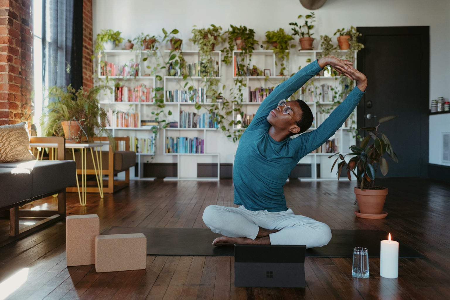 A man stretching on a yoga mat, a candle, a glass of water and boxes before him and bookshelves and plants behind him