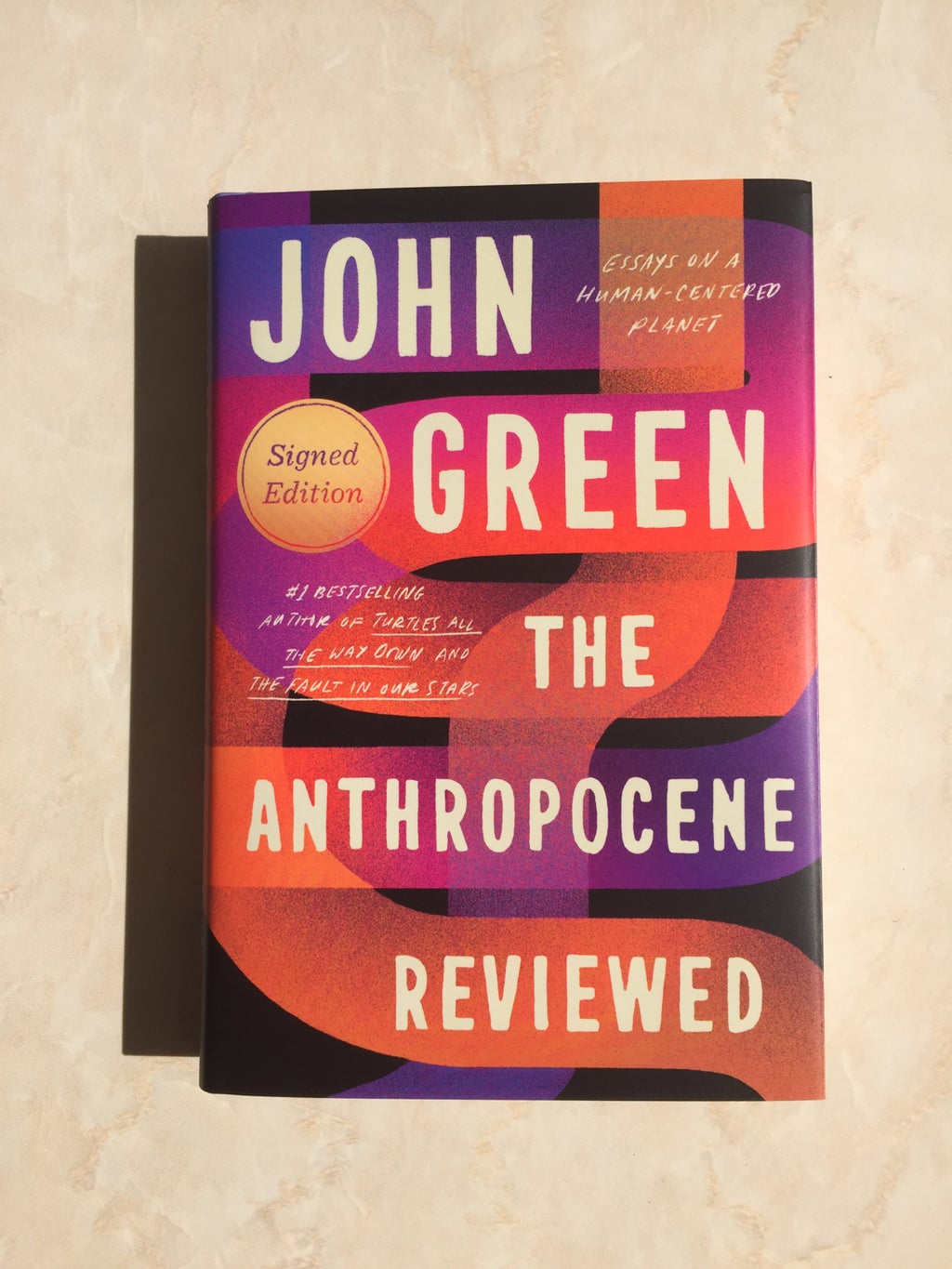 Picture of John Green\'s book \"The Anthropocene Reviewed\" against a plain backdrop