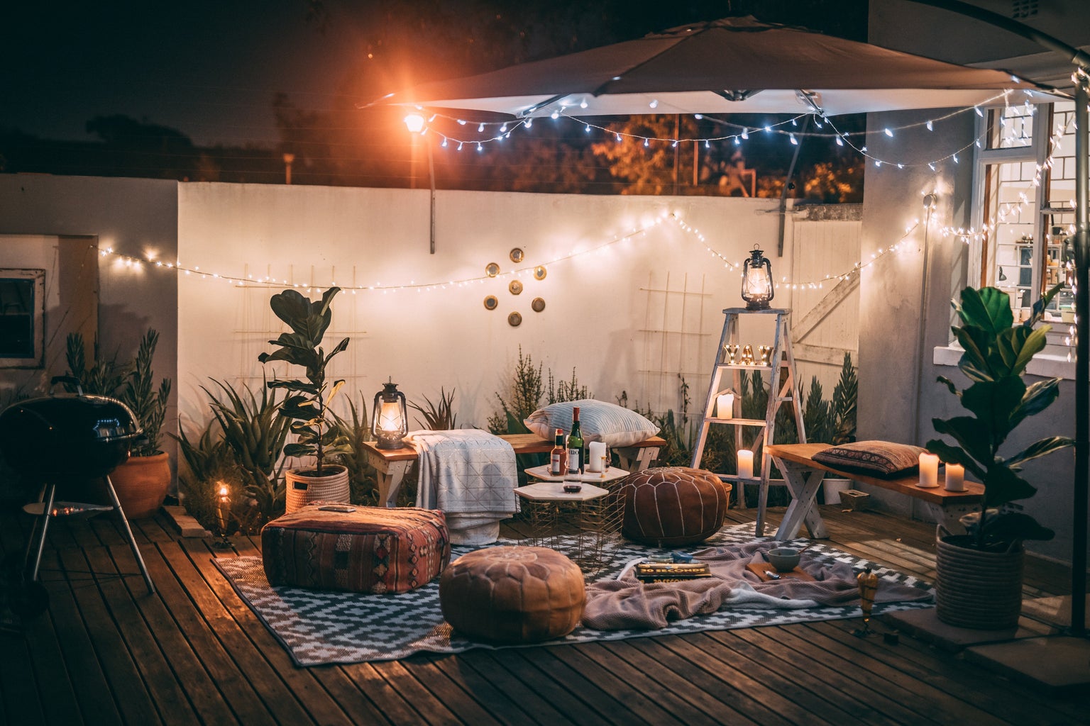 An outside setup with lots of cushions and some fairy lights. There are also plants everywhere and a small table that has drinks.