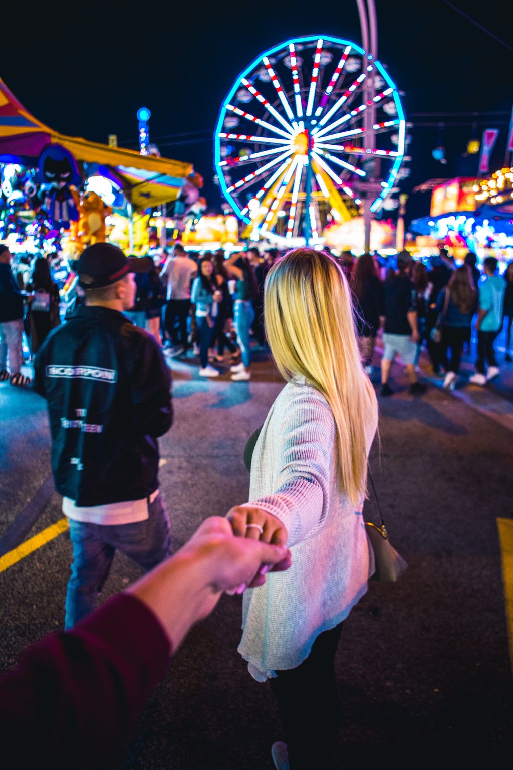 A woman holds a man\'s hands, facing away from the camera, and walks into a crowded fair.