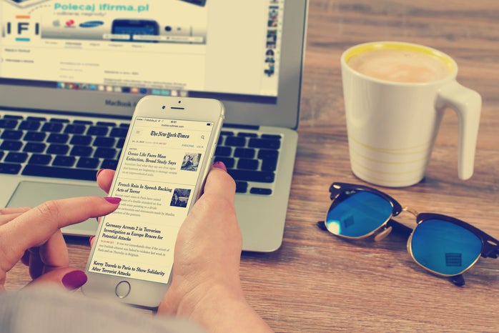 A person has their phone in their right hand. On the phone, The New York Times is seen. In the background, we see a laptop, a mug with what appears to be coffee and a pair of sunglasses.