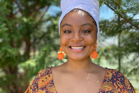 Girl is in a colorful dress and white headwrap smiling with trees in the background