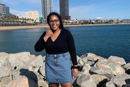 Young Black woman, Mercedes Owens, standing in front of the beach wearing a black top, jean skirt, and white shoes.