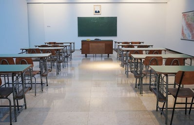 picture from the back of a classroom showing 10 empty desks and a green chalkboard in the center of the wall facing the classroom