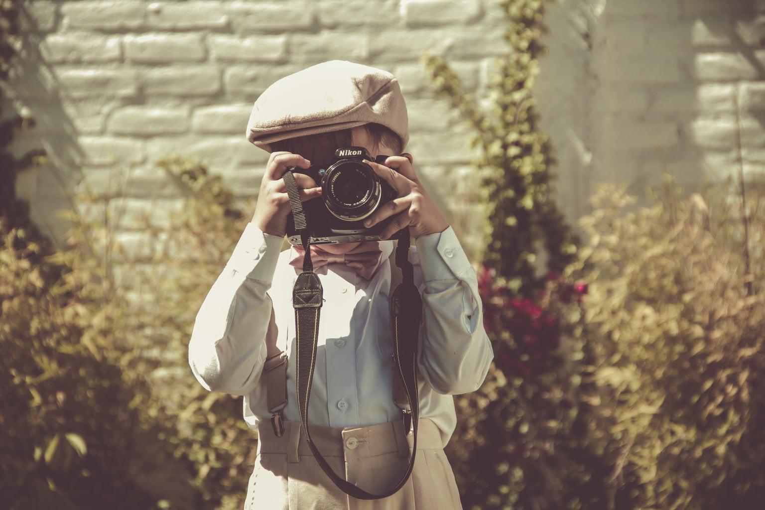 A boy in overalls and a beige hat holding up a Nikon and taking a photo