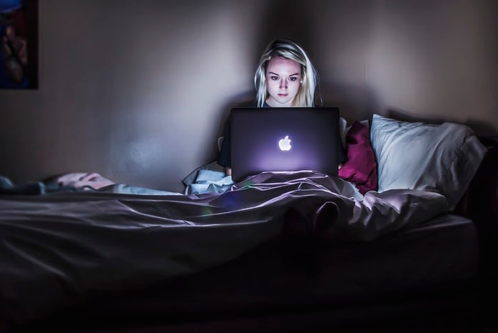 A young woman is sat in bed with a laptop in her lap. There is minimal lighting in the image except for the woman\'s face, which is illuminated by the laptop screen.