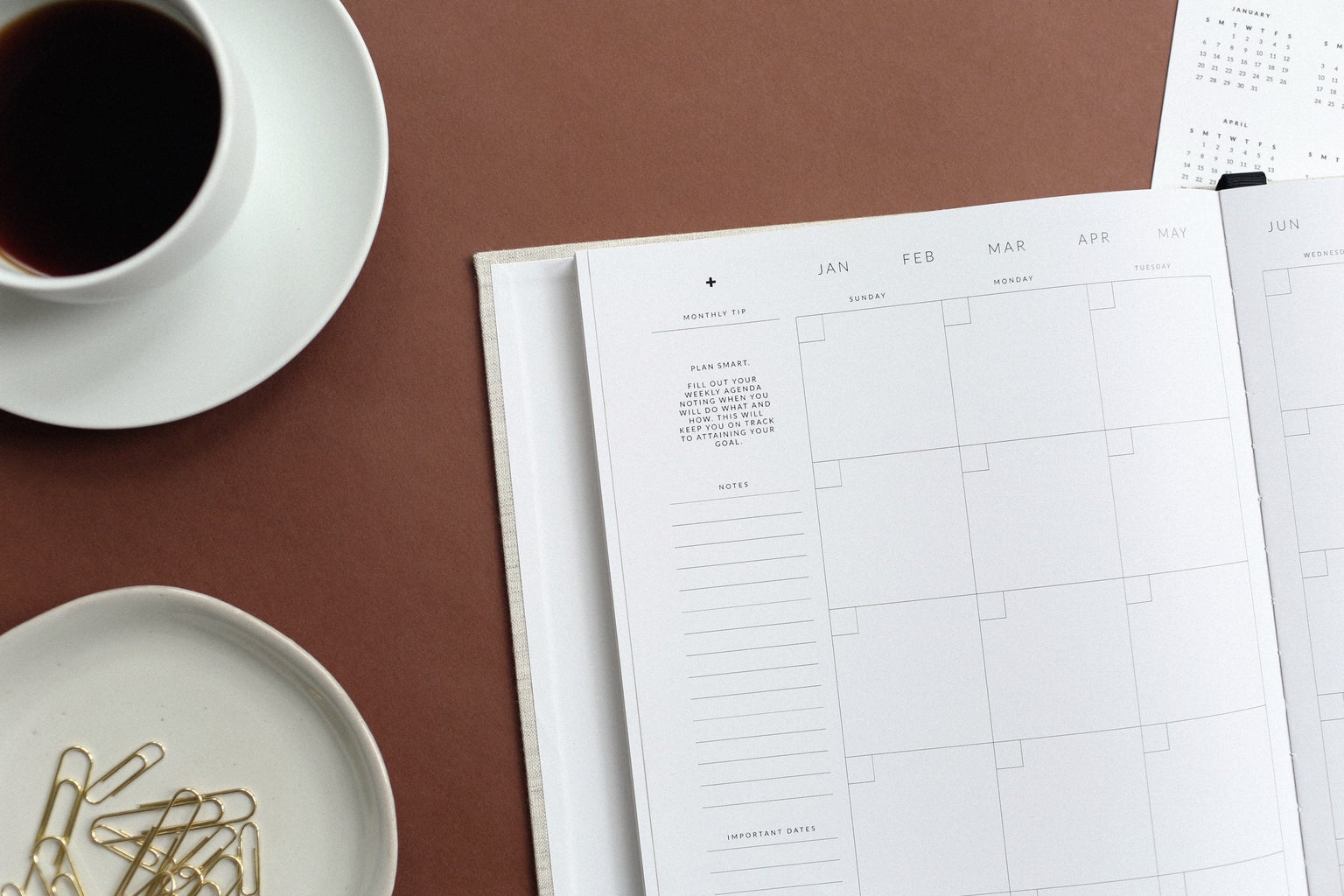 A blank open agenda on a table next to a full coffee mug on a plate and a plate filled with paperclips.