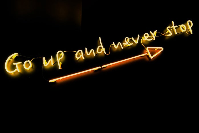 neon quote saying "go up and never stop" on a black background with an arrow underneath the words