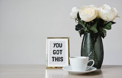 White flowers in a vase. There is a sign stating \"you got this\" to the left of the flowers and a teacup placed on top of a plate with a silverspoon placed inside.