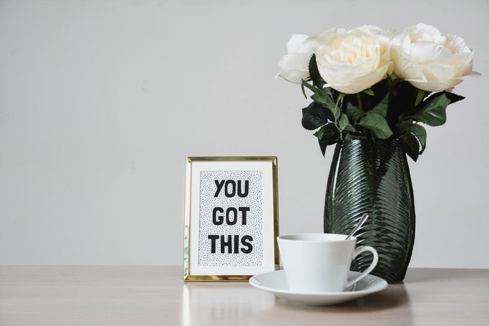 White flowers in a vase. There is a sign stating \"you got this\" to the left of the flowers and a teacup placed on top of a plate with a silverspoon placed inside.