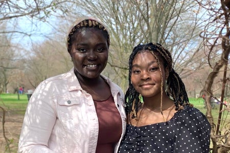 Two young Black women standing in a park together, one wearing a pink jacket and the other wearing a black and white polka dot top.