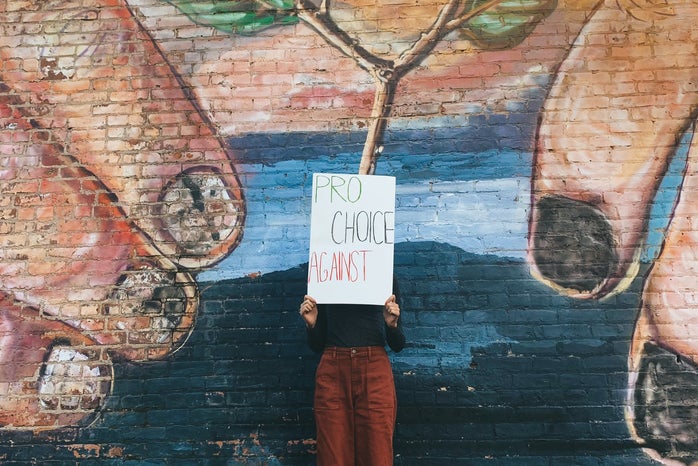 Woman standing in front of a wall with a mural and holding a sign that says \"Pro Choice Against\"