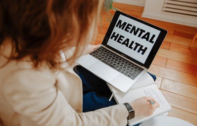 laptop open with \"mental health\" written on it while a woman writes in an open notebook