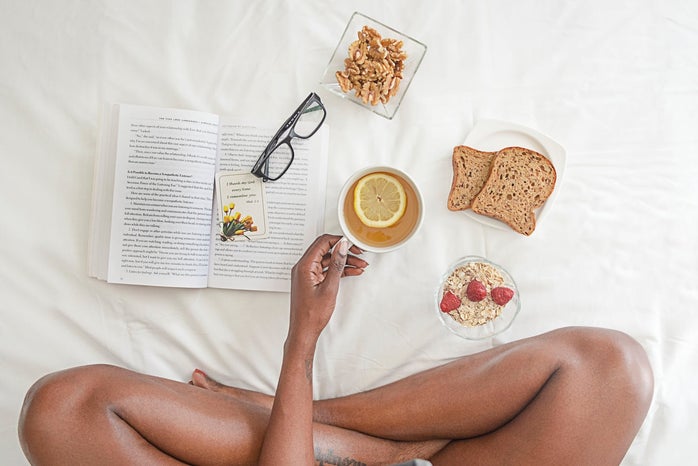Woman holding a white mug with breakfast food and a book open on a bed
