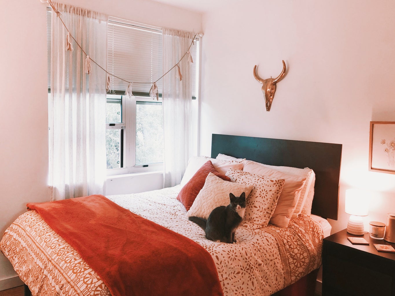 A bed that is decorated with burnt orange tones next to a window with sheer curtains