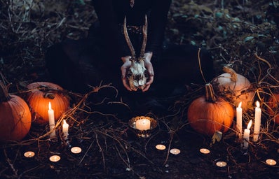 A witch sits among pumpkins and candles holding a jackalope skull