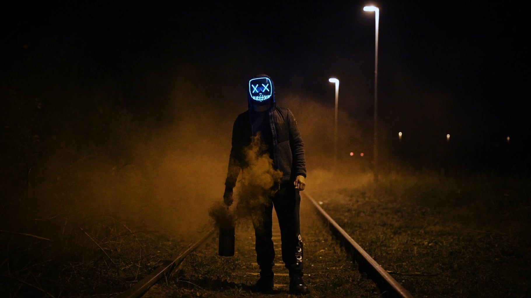 Figure stands on railway in a blue horror movie mask holding a smoking canister.