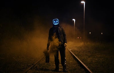 Figure stands on railway in a blue horror movie mask holding a smoking canister.