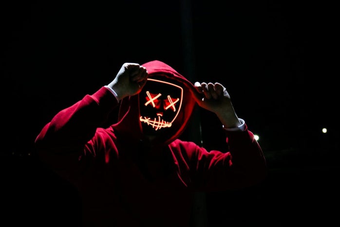 A person in a red hoodie holds his hands to the hood, and is wearing a red horror movie mask.