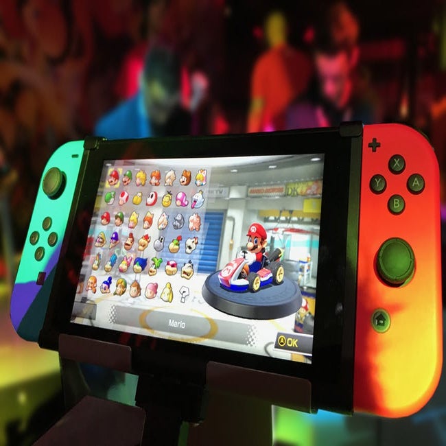 A nintendo switch displaying mario kart is on a display case in front of  blurry convention scene in the background