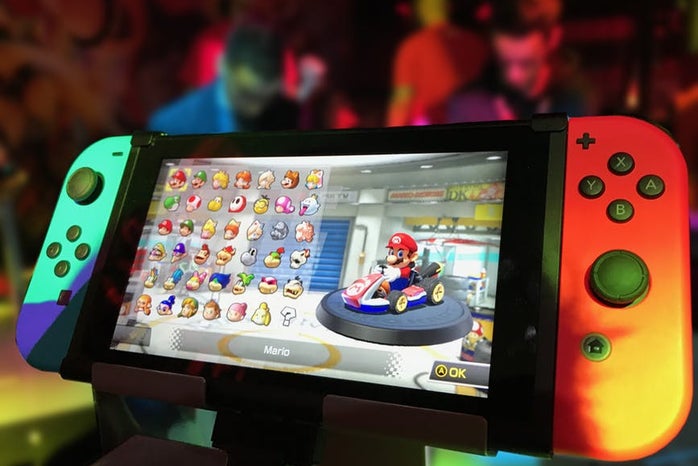 A nintendo switch displaying mario kart is on a display case in front of  blurry convention scene in the background