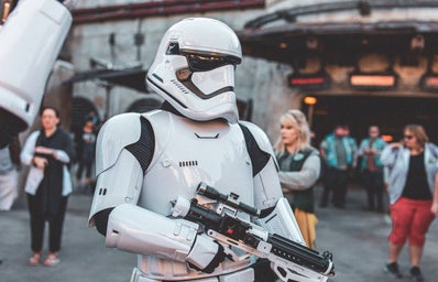 A Star Wars Stormtrooper stands in front of an out-of-focus Galaxy\'s Edge in Disney World. The entire image has a cool filter.