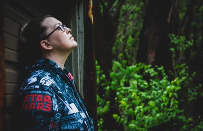 A female star wars fan in glasses leans against a wall near some trees