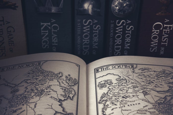 A book is open to pages of drawn maps, the background is a shelf containing the Game of Thrones books