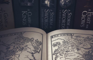 A book is open to pages of drawn maps, the background is a shelf containing the Game of Thrones books