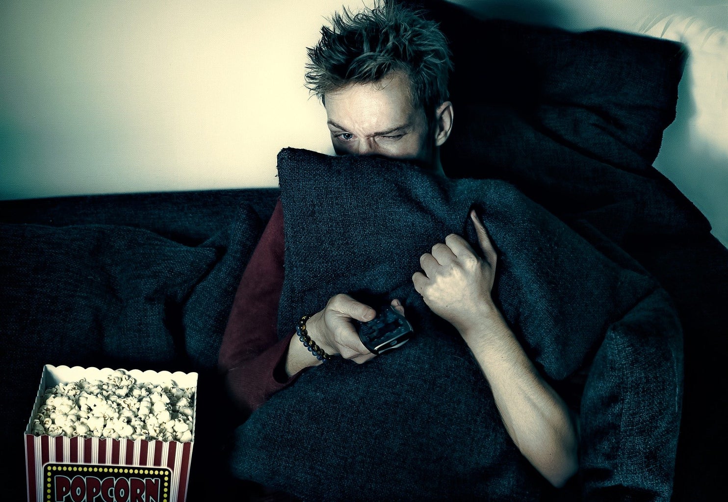 A man is sitting on a couch, next to popcorn in the dark, hiding behind a pillow