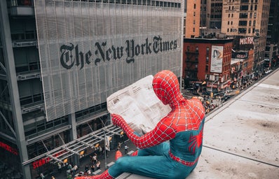 A man in a spiderman costume reads the newspaper on top of a building