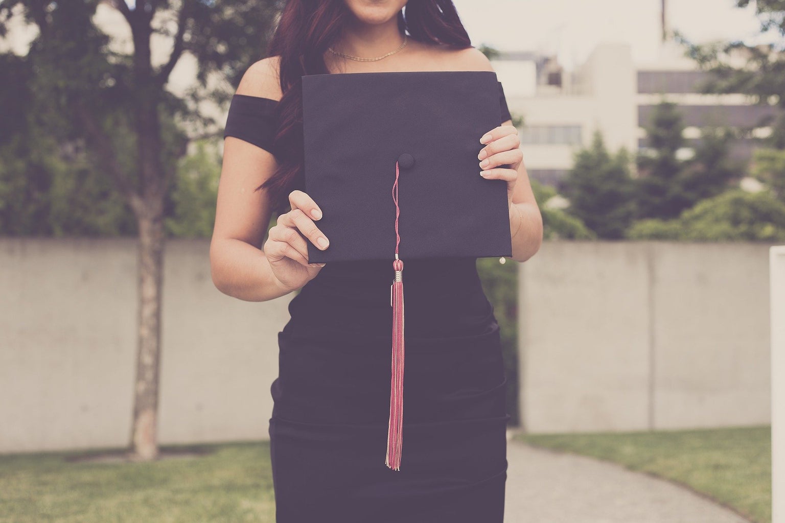 woman in black dress stands outside in front of a fence holding her graduation cap