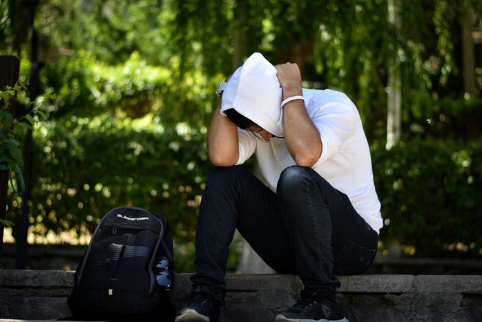 a man sits on outdoor steps in a white hoodie with his hands behind his neck looking frustrated