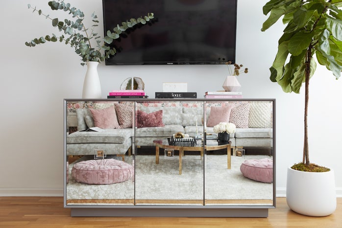 a mirrored cabinet under a mounted television with plants and books to accent. the couch and coffee table are reflected.